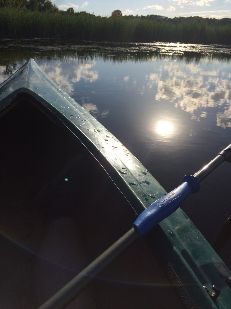 Sunlight and white, puffy clouds reflect upon a marsh's waters beside the nose of a green kayak